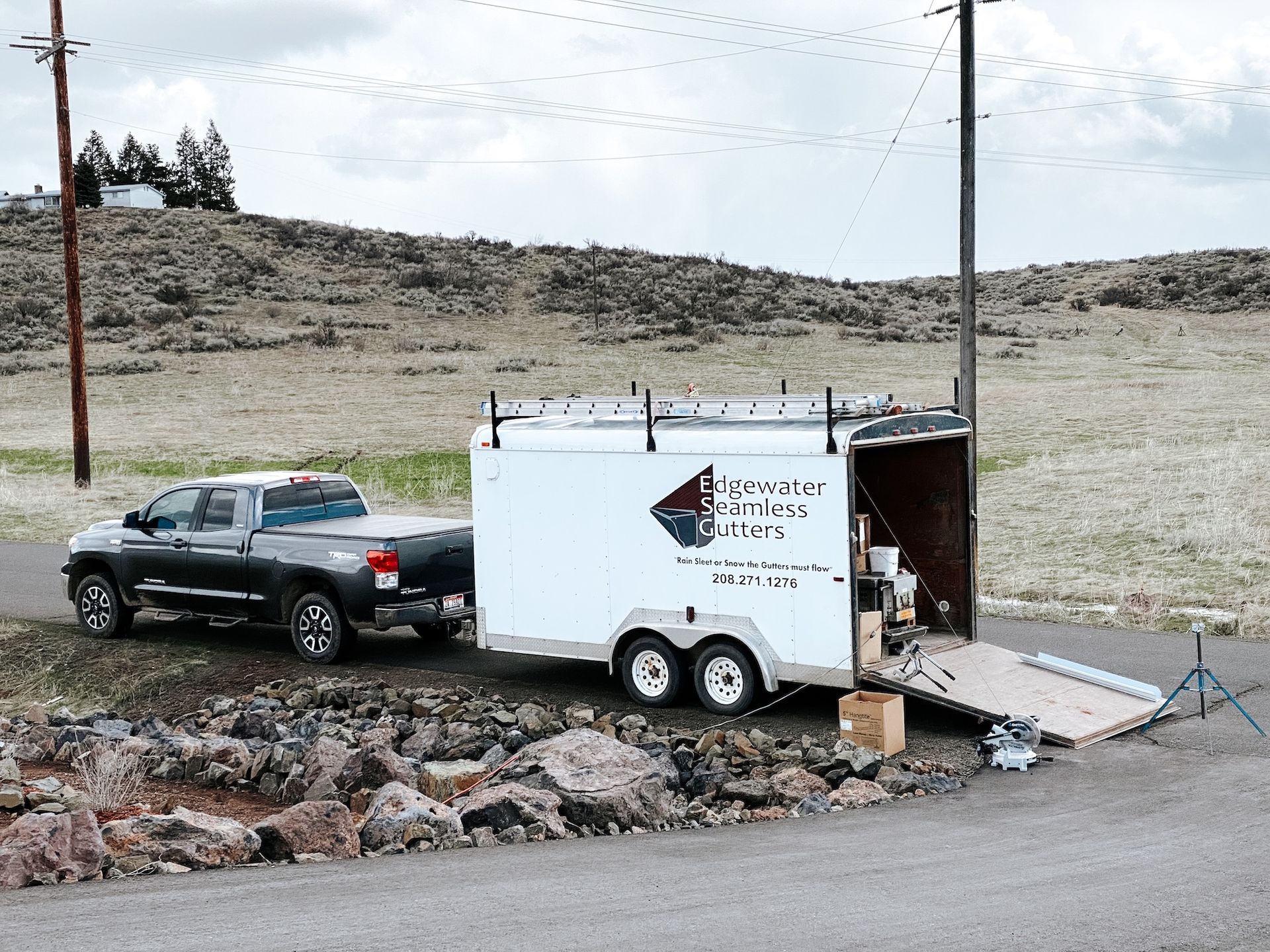 Black pickup towing a white utility trailer parked on a dirt road in a rocky, rural area.