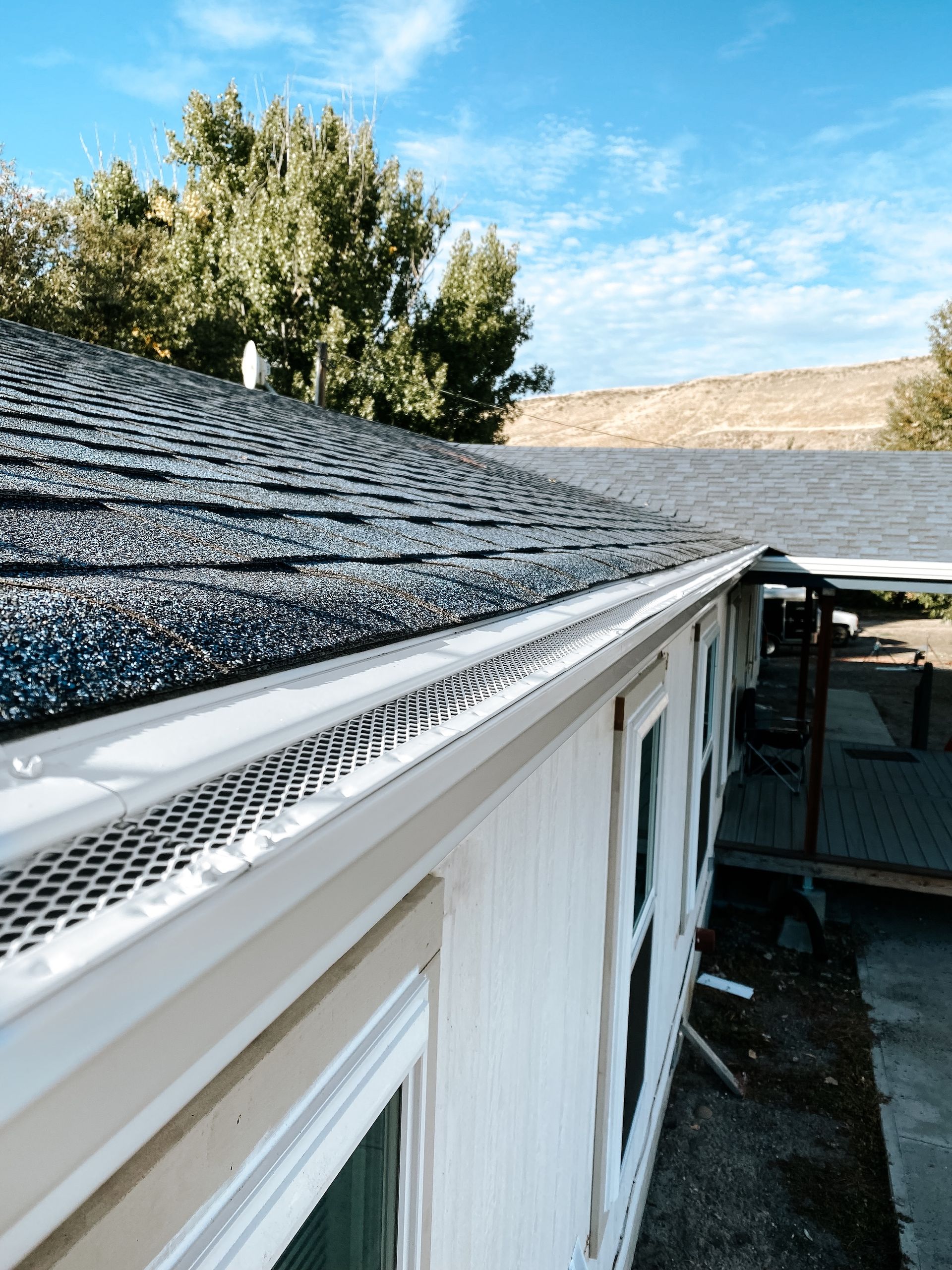 Roof gutter along white house exterior with dark shingle roof and tree under blue sky
