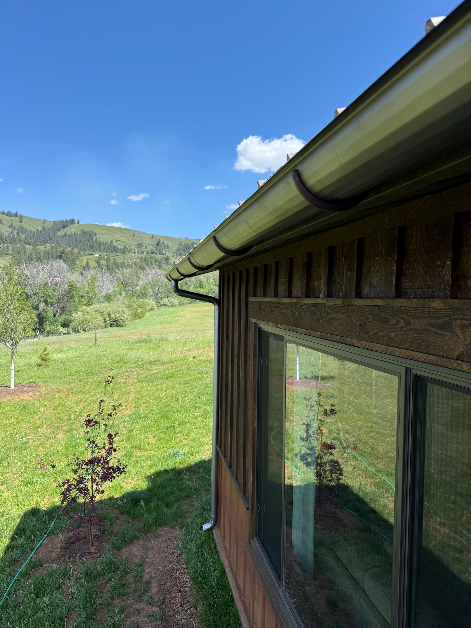 Mountain cabin with large windows beside a green meadow and forested hills under a blue sky