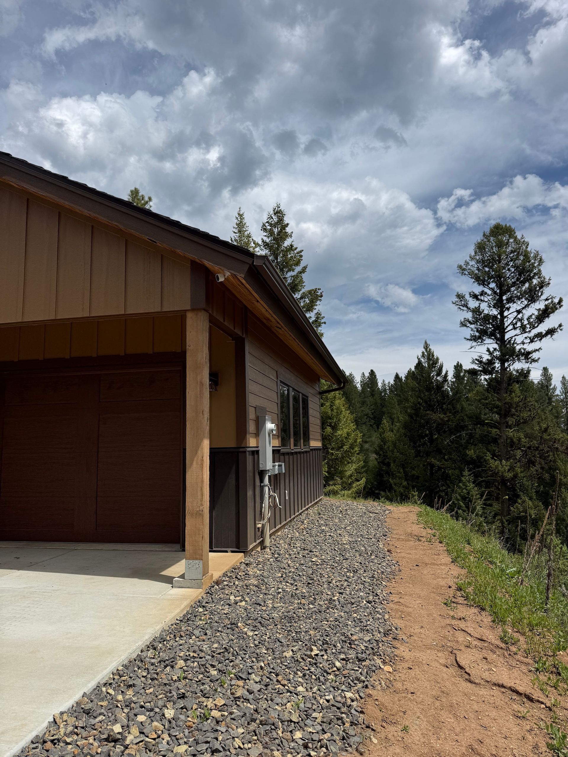 House exterior with gravel drainage bed, concrete driveway, and cloudy sky beside pine trees