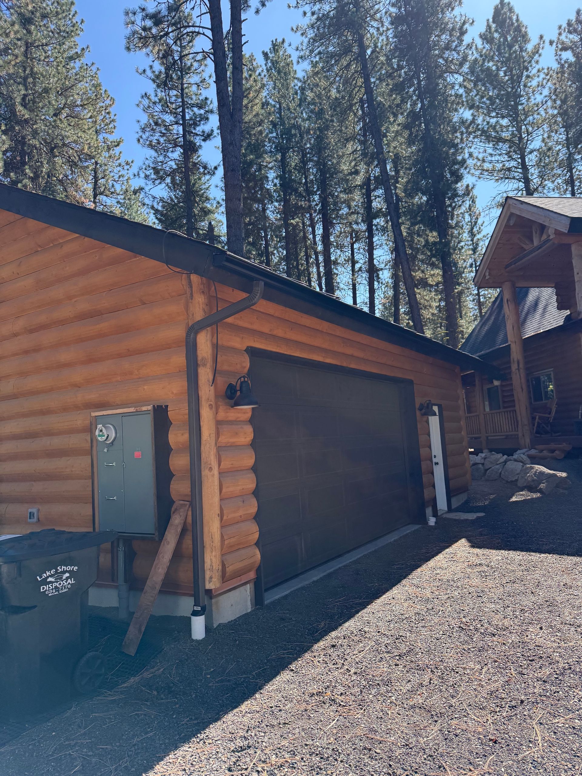 Log cabin garage with brown door beside gravel driveway and pine trees under a clear sky
