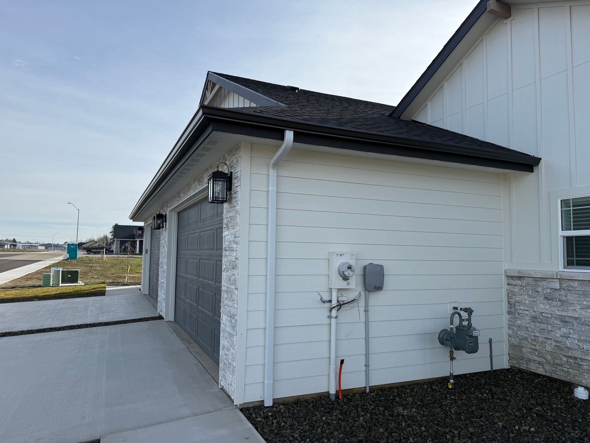 Side view of a white house exterior with utility meters, downspout, and concrete walkway by a road