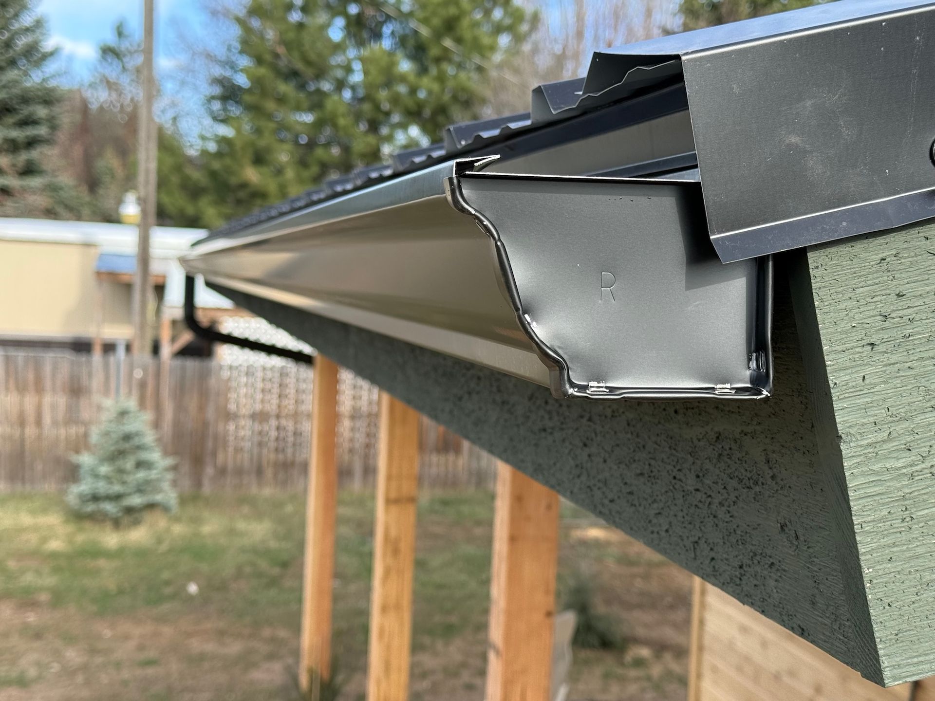 Close-up of a black metal gutter and roof edge on a house, with backyard fencing and trees behind.
