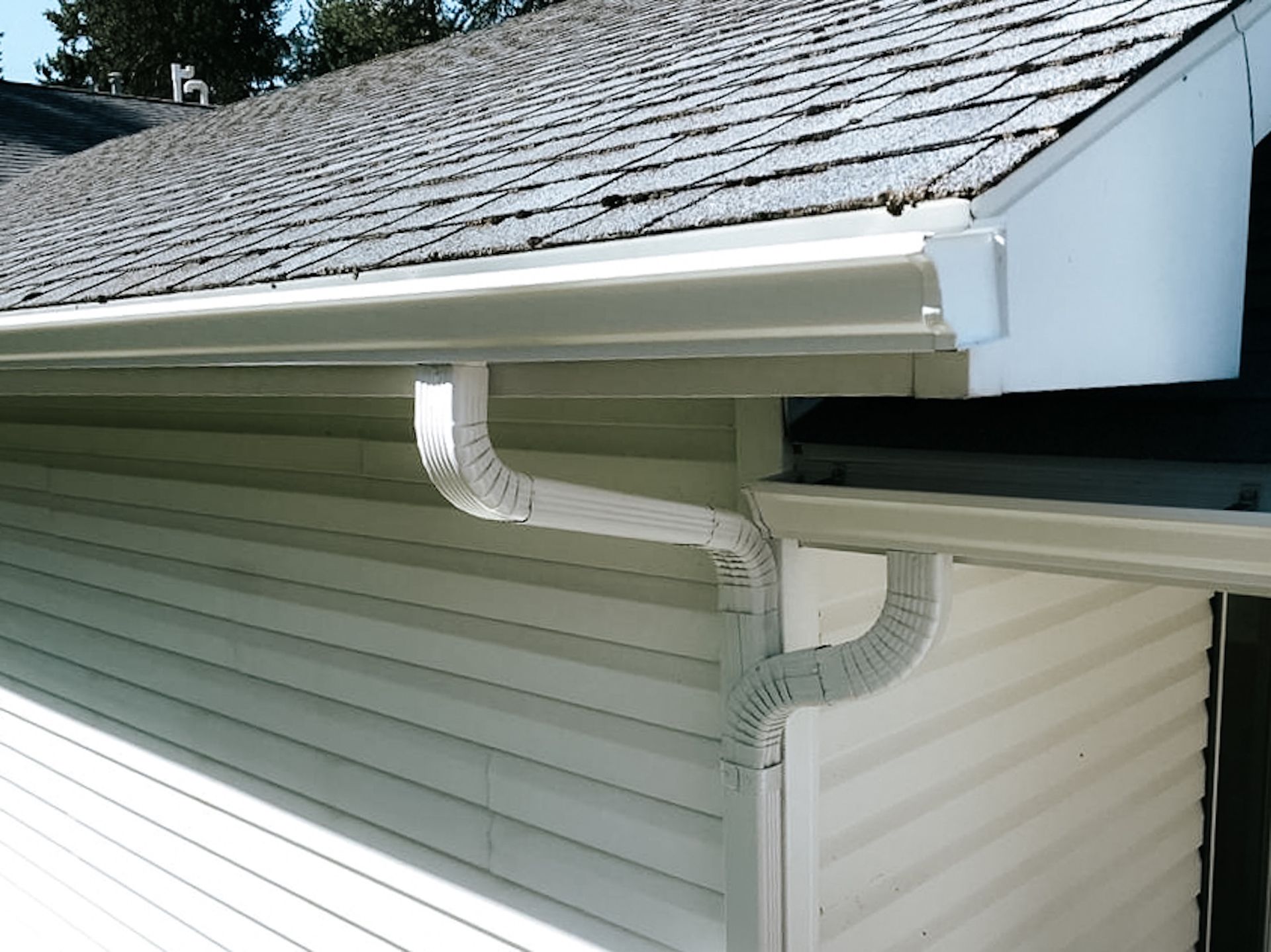 White house gutter and downspout attached to a vinyl-sided roofline, with a clean eave corner
