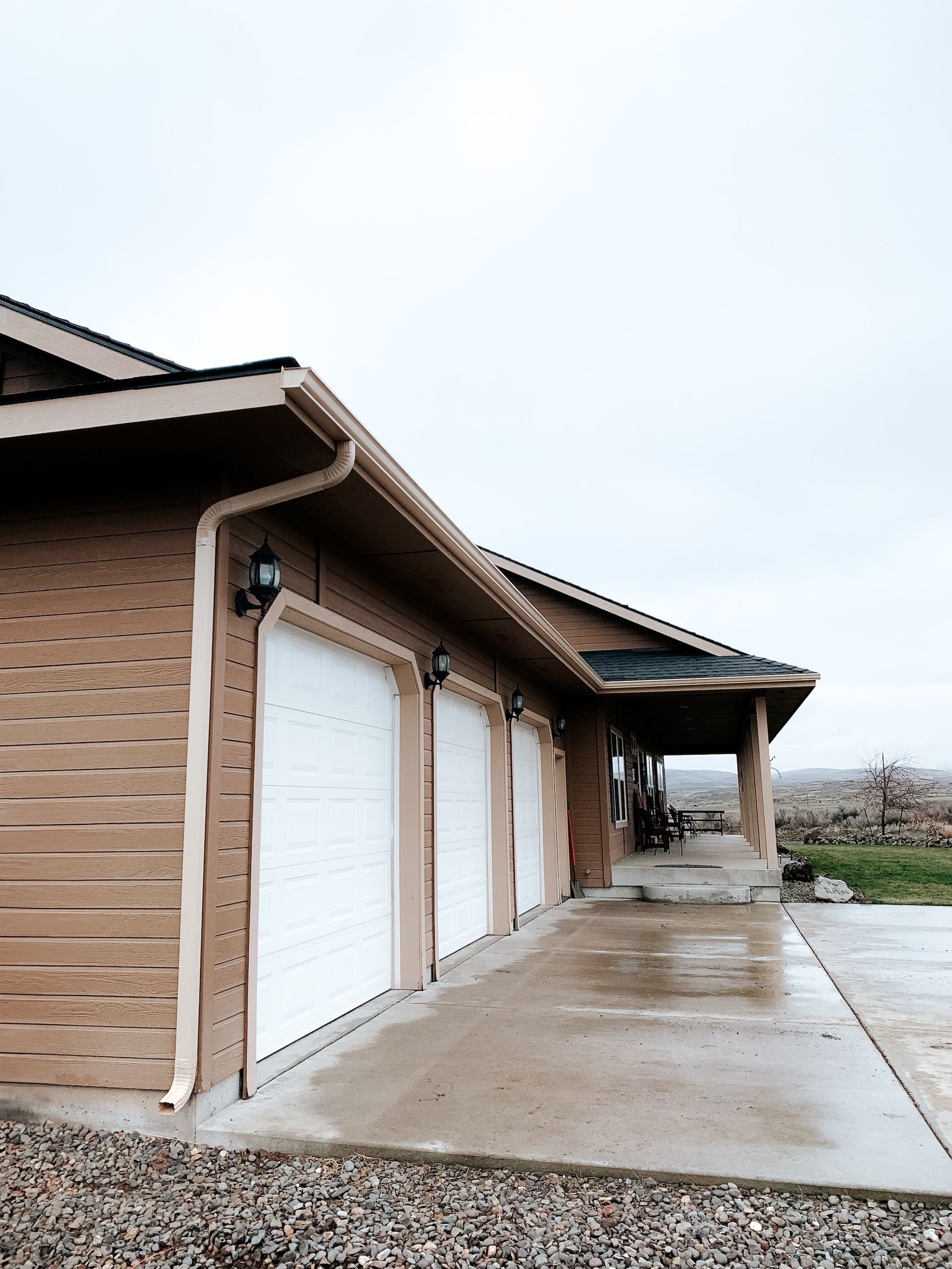 Row of tan garage units with white doors beside a wet concrete driveway under an overcast sky