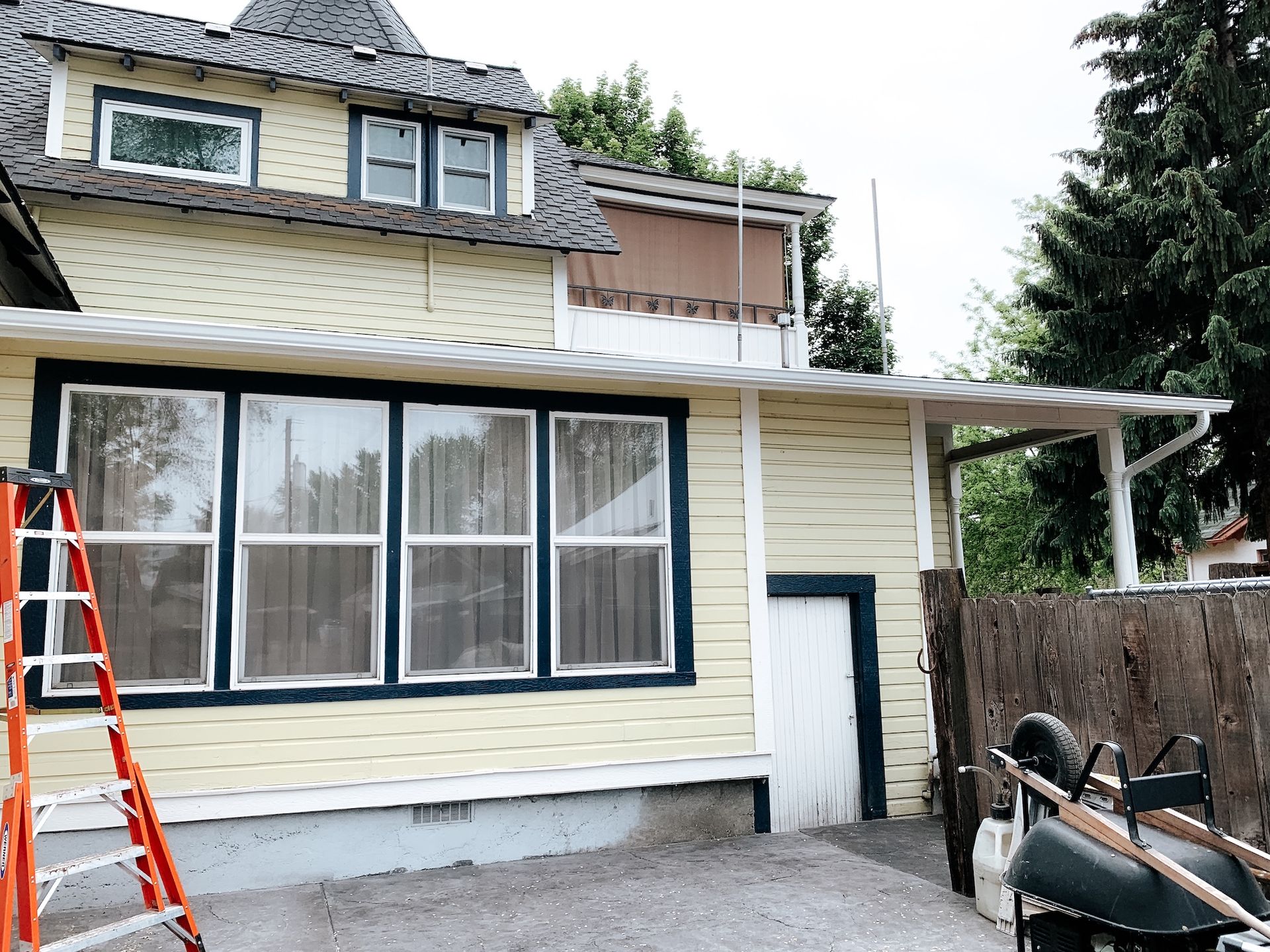 Exterior of a house with cream siding, large front windows, a red ladder, and construction materials in the yard