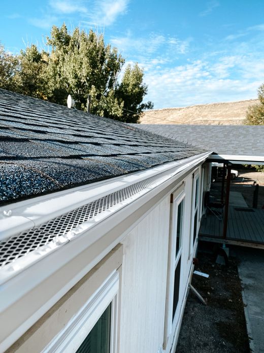 Roof gutter along a house exterior beside a dark shingled roof under a blue sky