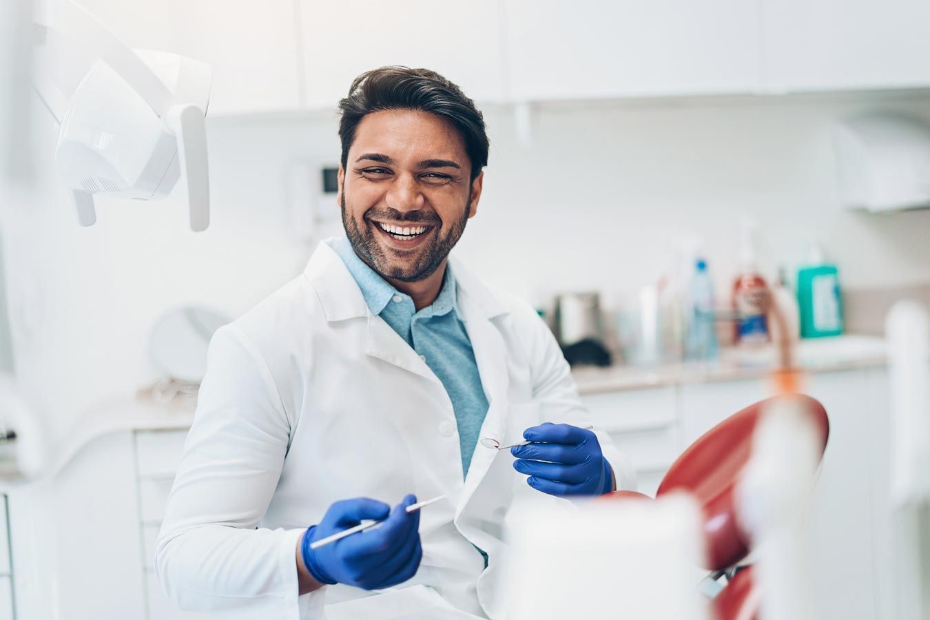 Dentist smiling in a bright clinic, wearing a white coat and blue gloves while holding dental tools