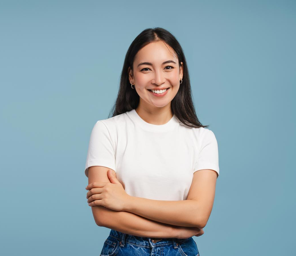 Smiling woman with crossed arms, wearing a white shirt and blue jeans against a light blue background