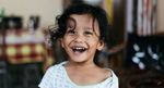 Smiling child with messy hair indoors, wearing a white shirt, in a softly blurred room