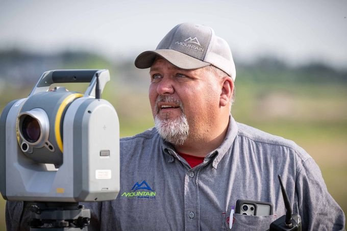 Man in cap operates surveying equipment outdoors, looking right.