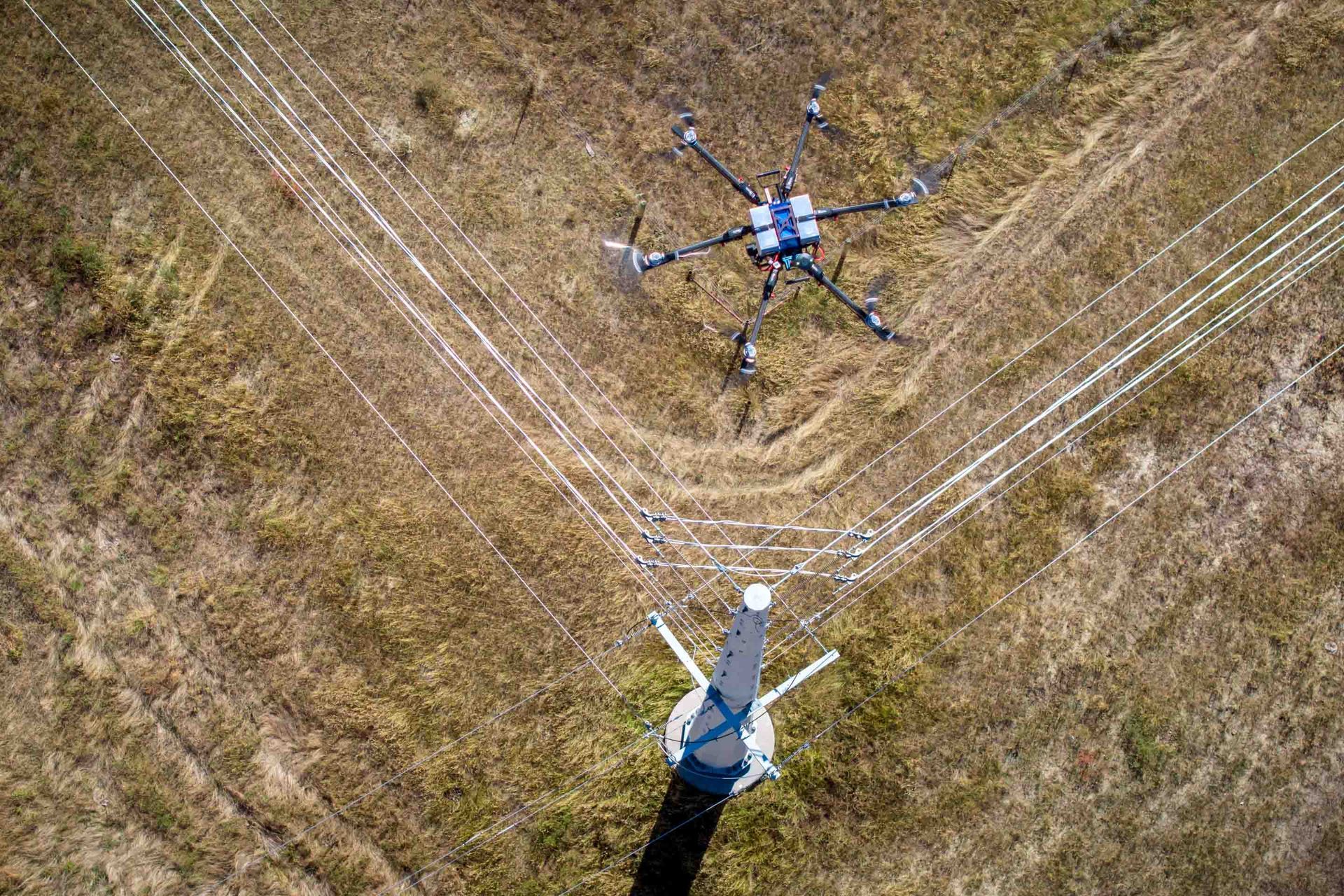 Drone inspecting power lines in a field. Top-down view with lines and a tower visible.