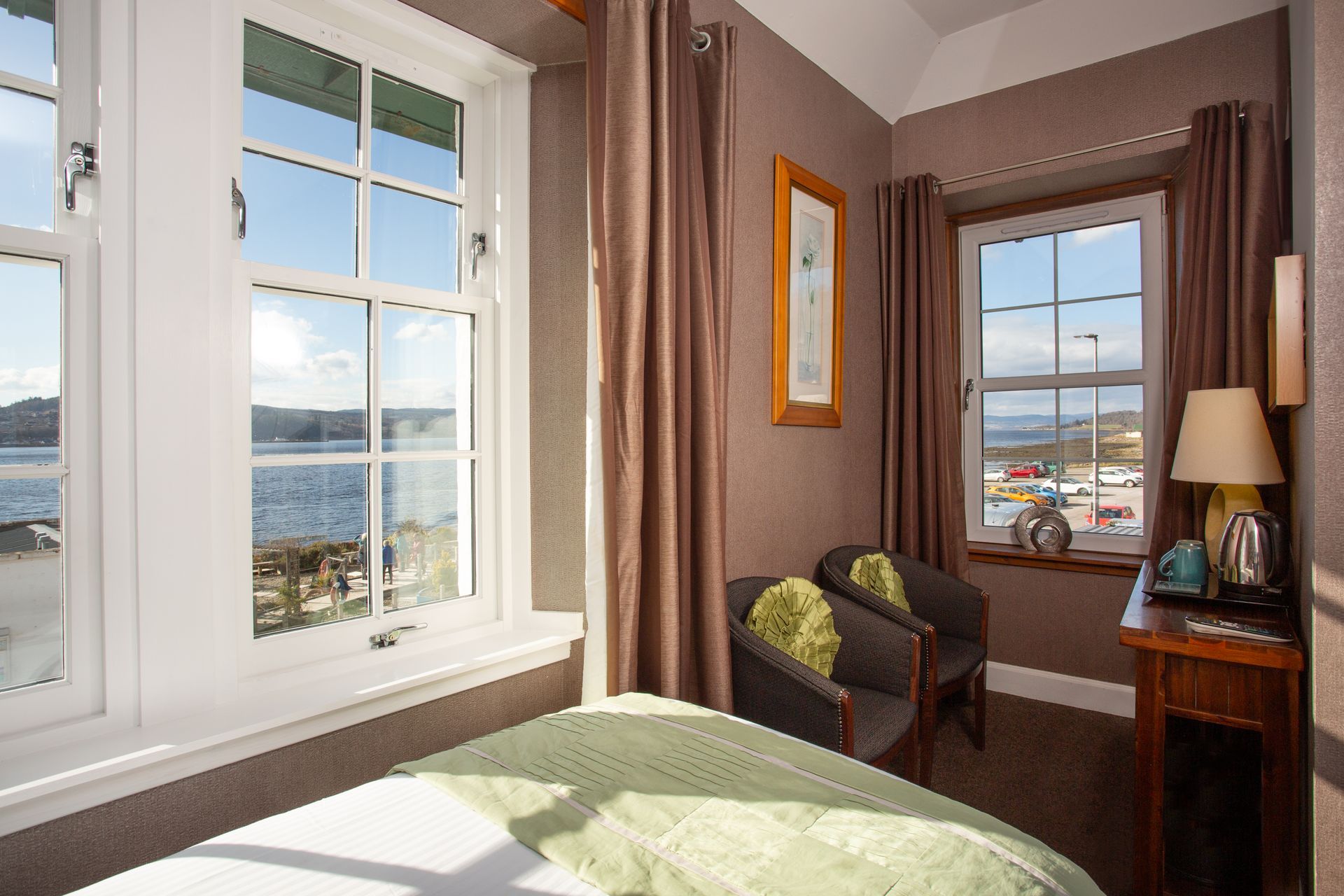 A hotel room with a bed , chairs , desk and two windows.