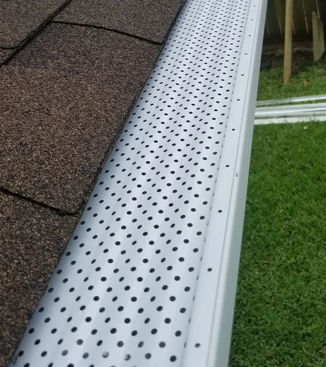 Gutter guard with small holes installed on a brown shingle roof, over a white gutter.
