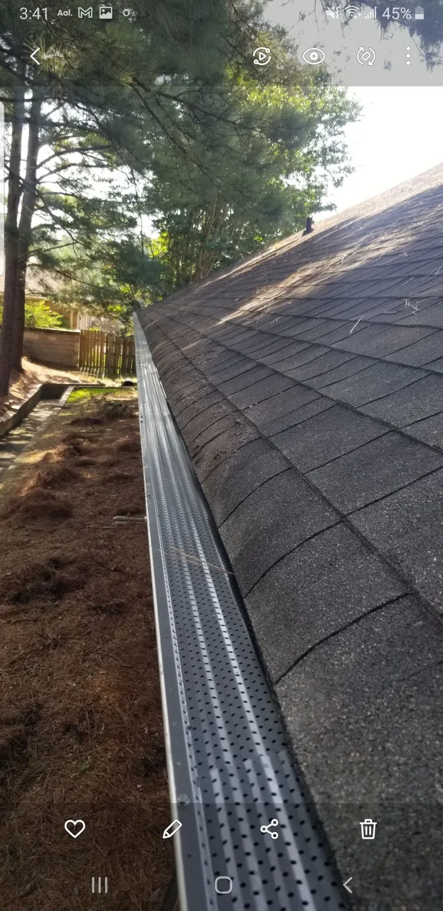 A roof with a gutter lined with a metal mesh cover. Brown shingles and a background of trees.