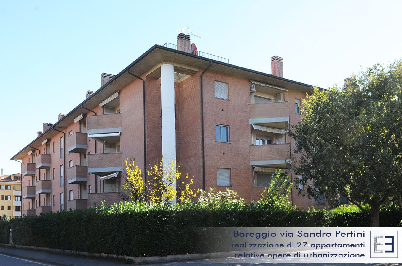 Palazzo di appartamenti in mattoni con balconi e siepi lungo una strada soleggiata, con un'insegna sulla facciata.