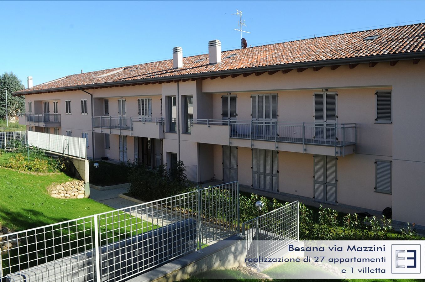 Palazzo residenziale con balconi e tetto di tegole, visto da un cortile erboso sotto un cielo azzurro e limpido.