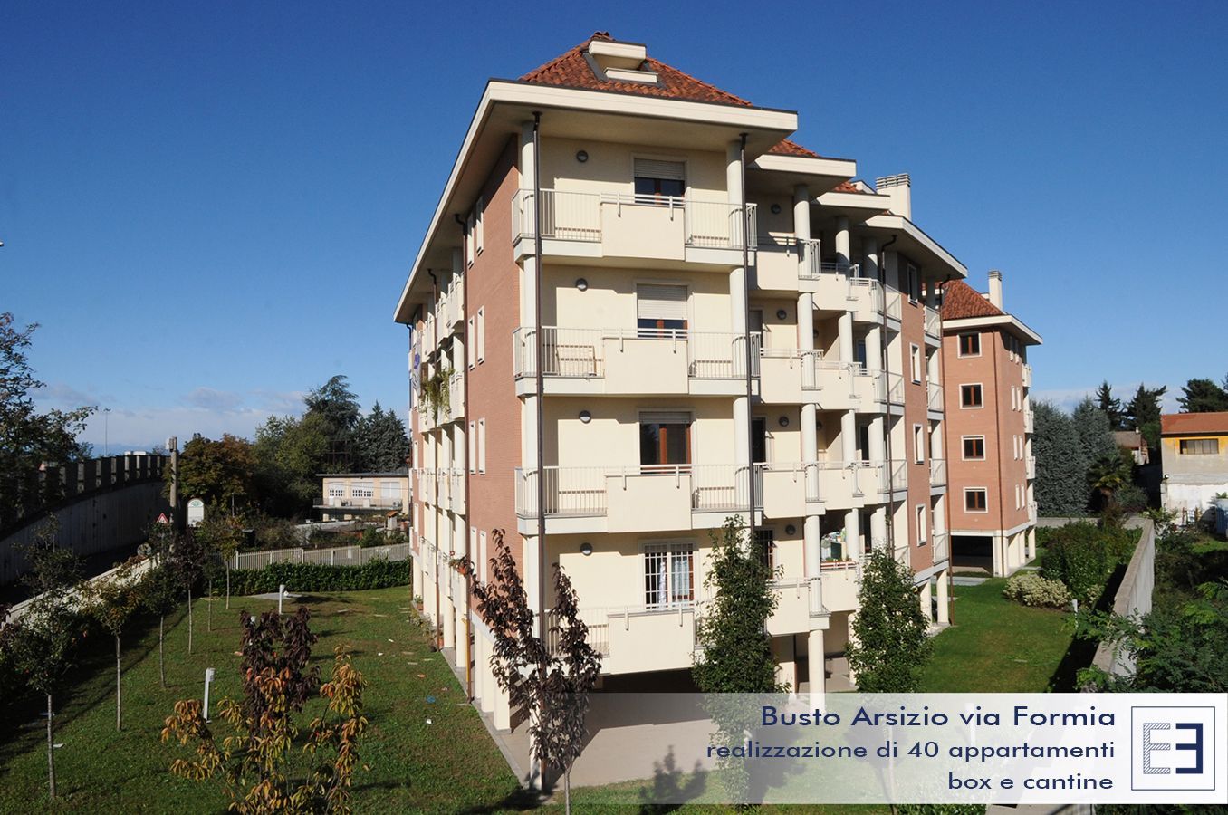 Palazzo residenziale con balconi beige e tetto rosso, affacciato su un sentiero erboso sotto un cielo azzurro.