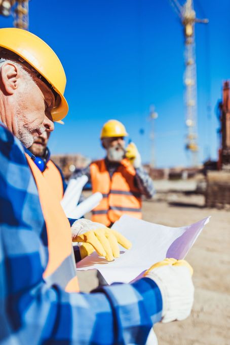 Operai edili con elmetti di sicurezza esaminano i progetti in un cantiere con gru sullo sfondo.