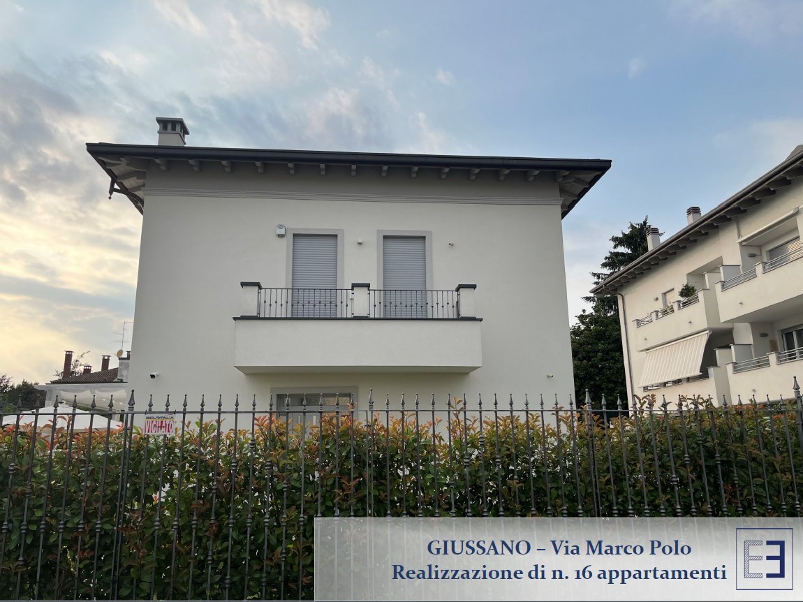 Casa bianca a due piani dietro una siepe al crepuscolo, con balcone e persiane chiuse.