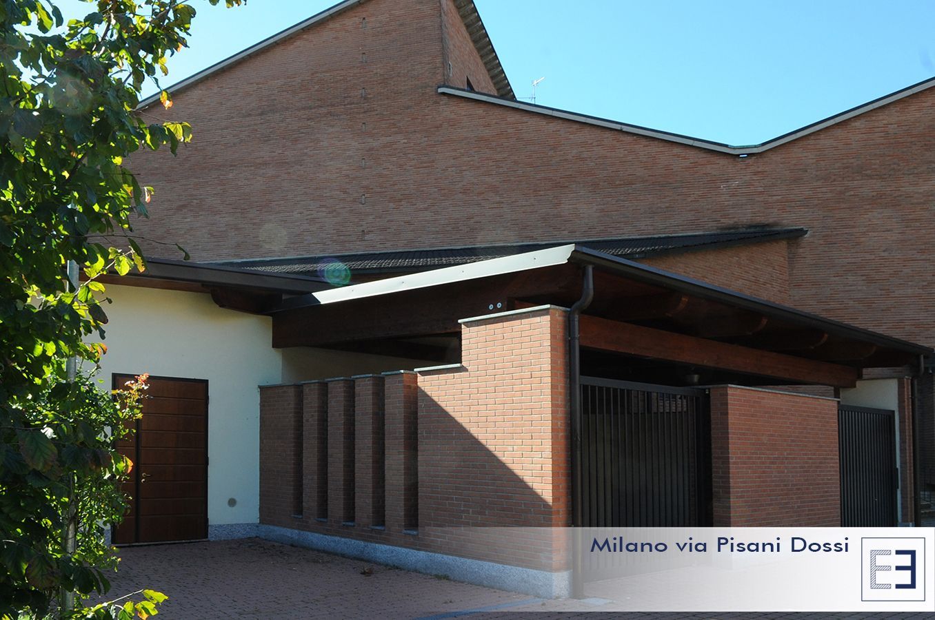 Ingresso di un edificio in mattoni con tetto spiovente, pareti bianche e alberi che proiettano ombre.