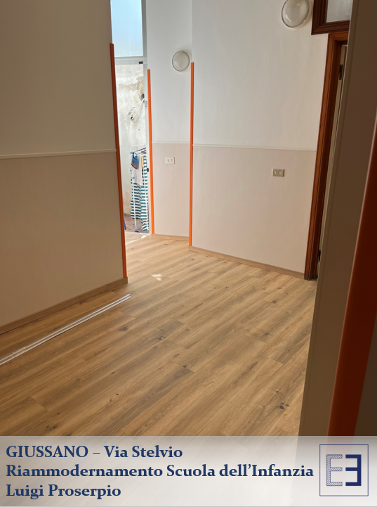 Corridoio vuoto con pavimento in legno chiaro, pareti bianche, porta aperta e vista sull'ingresso di una scuola.