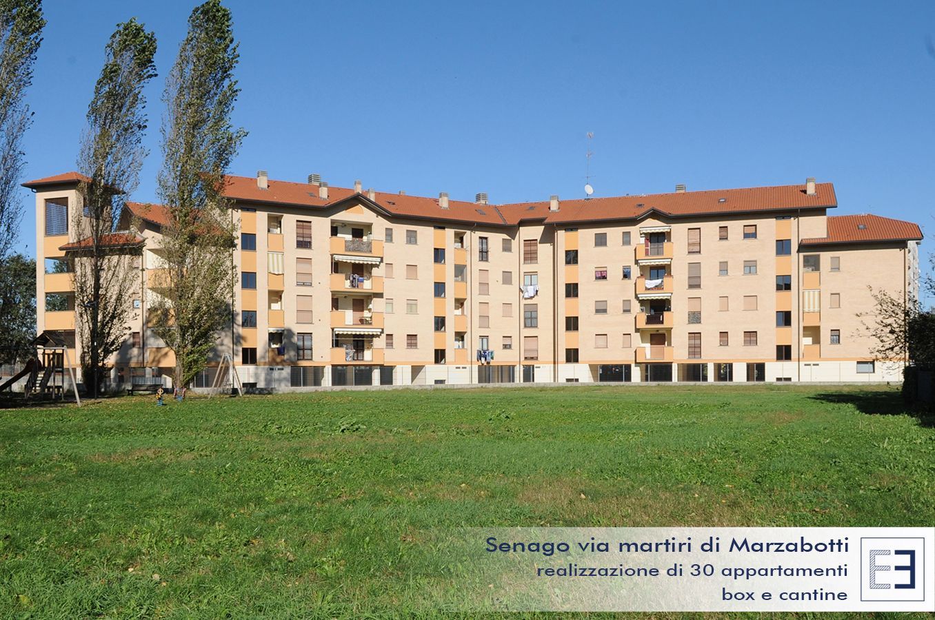 Palazzo di appartamenti con tetto rosso, sullo sfondo di un prato verde e alberi ad alto fusto, sotto un cielo limpido.