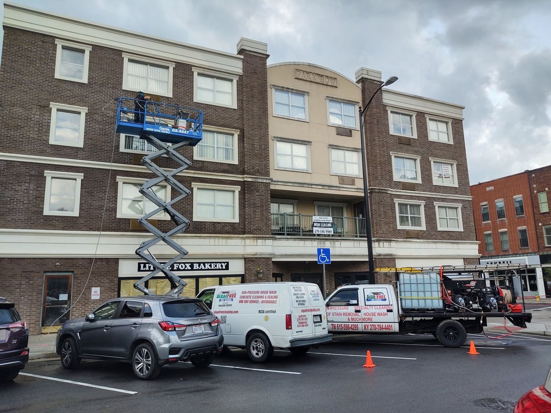 Workers on a lift inspecting a brick apartment building, with parked vans and a car below.
