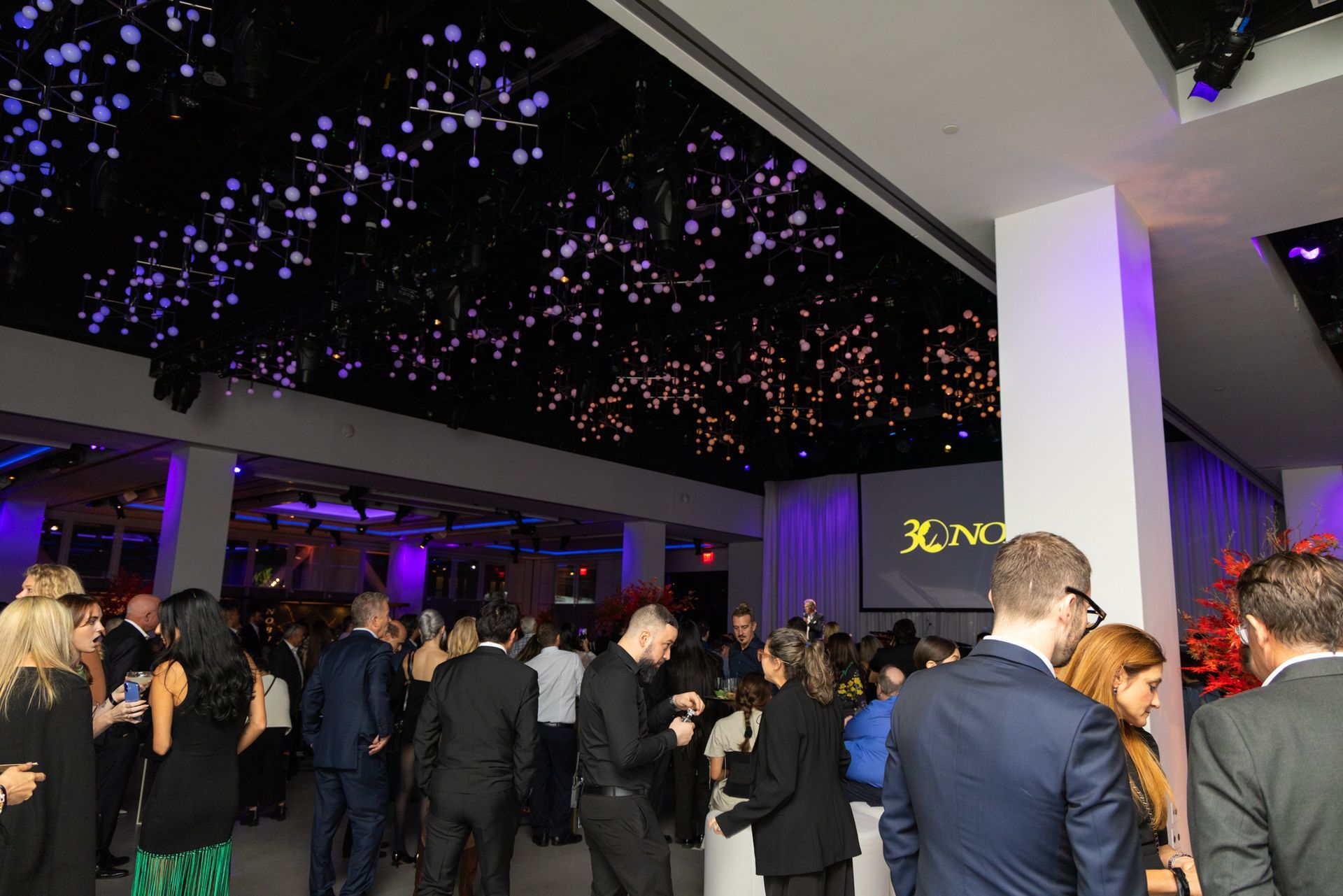Large event space with many people mingling; decorative ceiling lights.