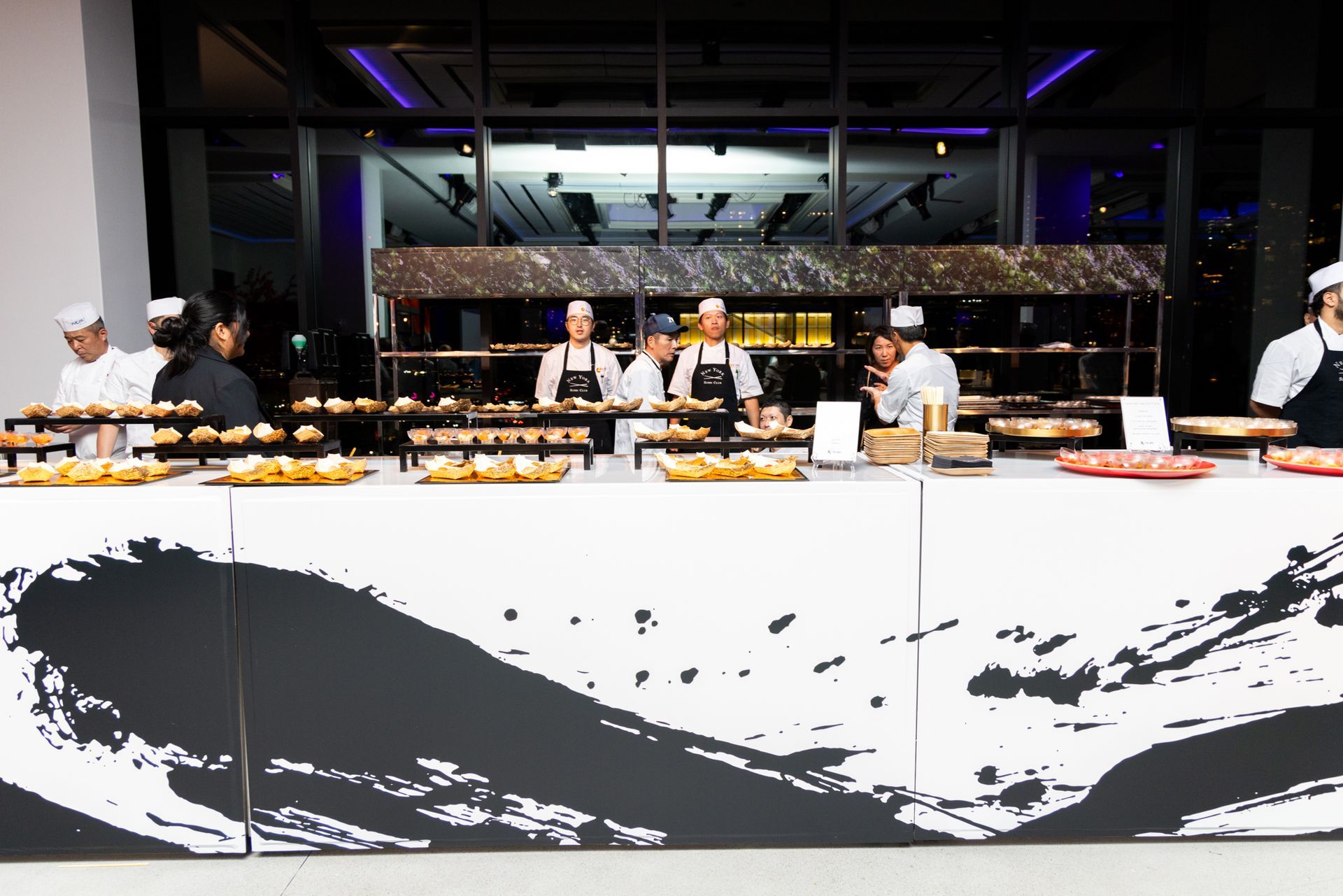 Chefs at a buffet table with various foods. The table has black and white art. A building with windows is in the background.