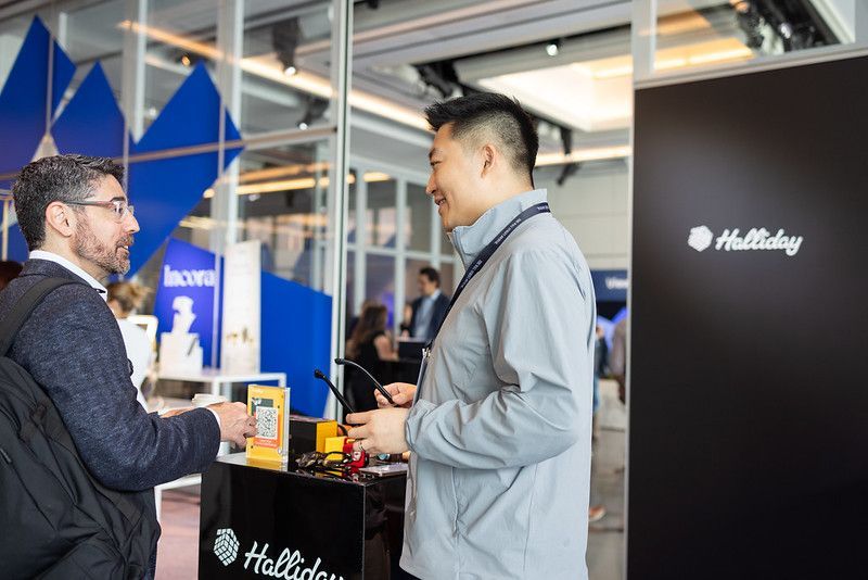 Two men conversing at a trade show booth with the Halliday logo. The setting is indoors, with a modern design.