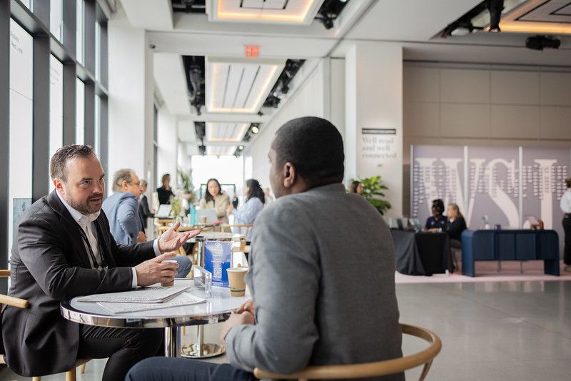 Two men converse at a table in a modern, open space with others in the background.