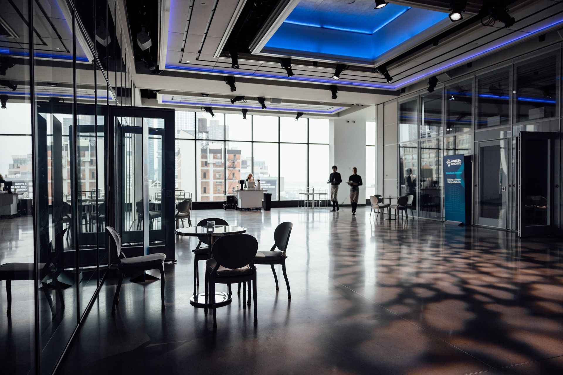 Large, modern room with floor-to-ceiling windows and reflective wooden floors. Chairs and tables are set up.
