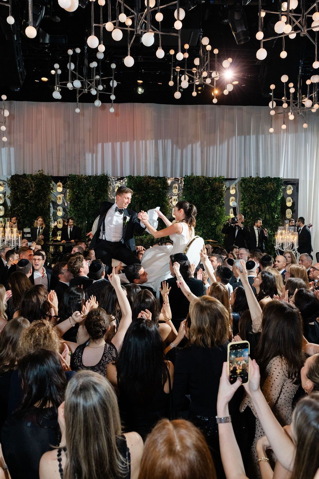 Couple being lifted and danced on chairs at a wedding reception. People in formal attire cheering.