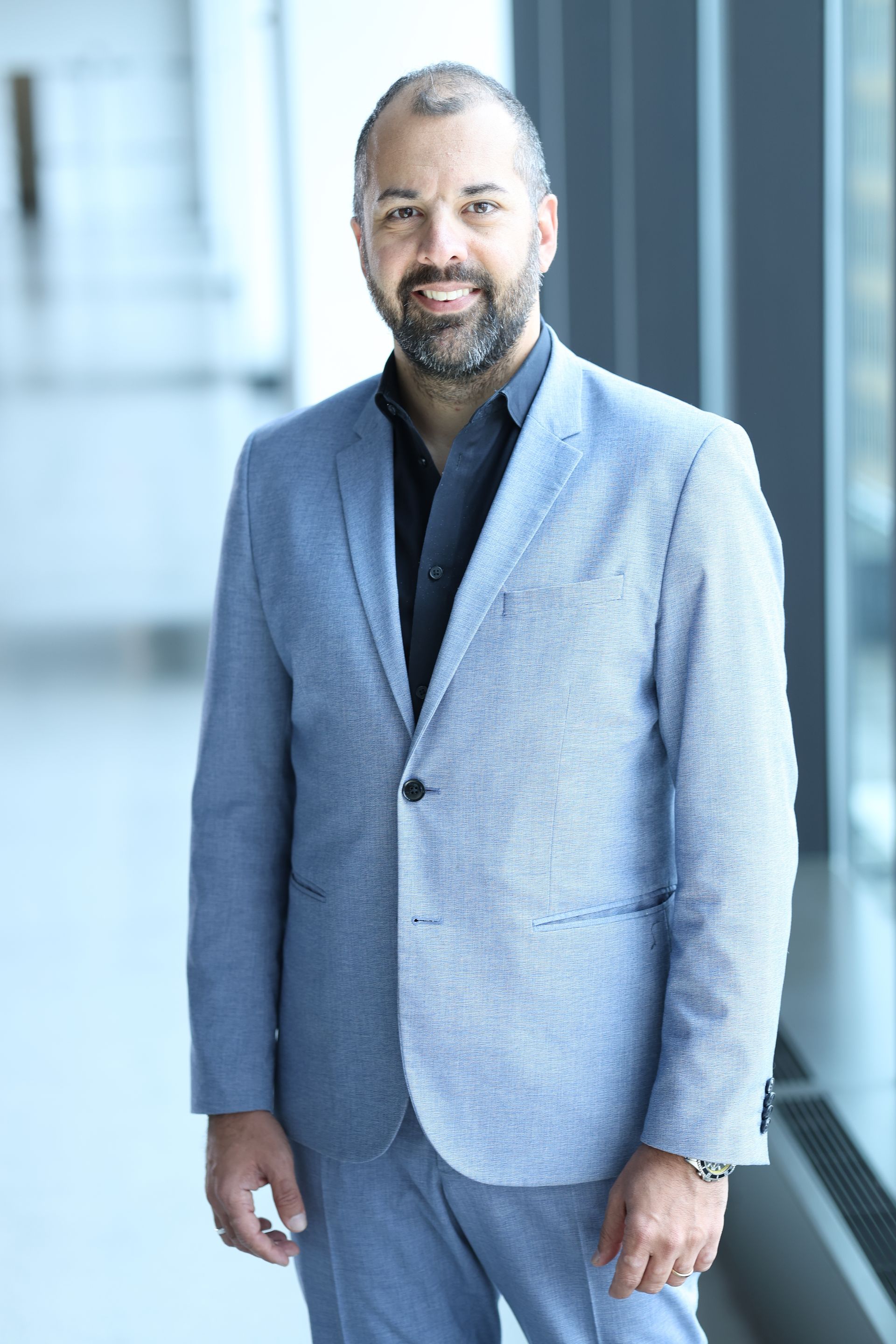 Man in light blue suit, black shirt, smiling, standing near a window.