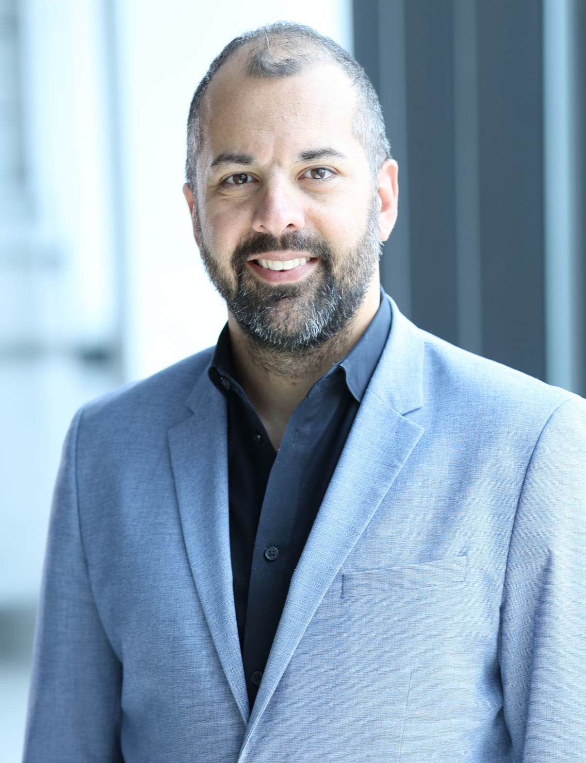 Man with a beard wearing a light blue blazer and black shirt, smiling indoors.