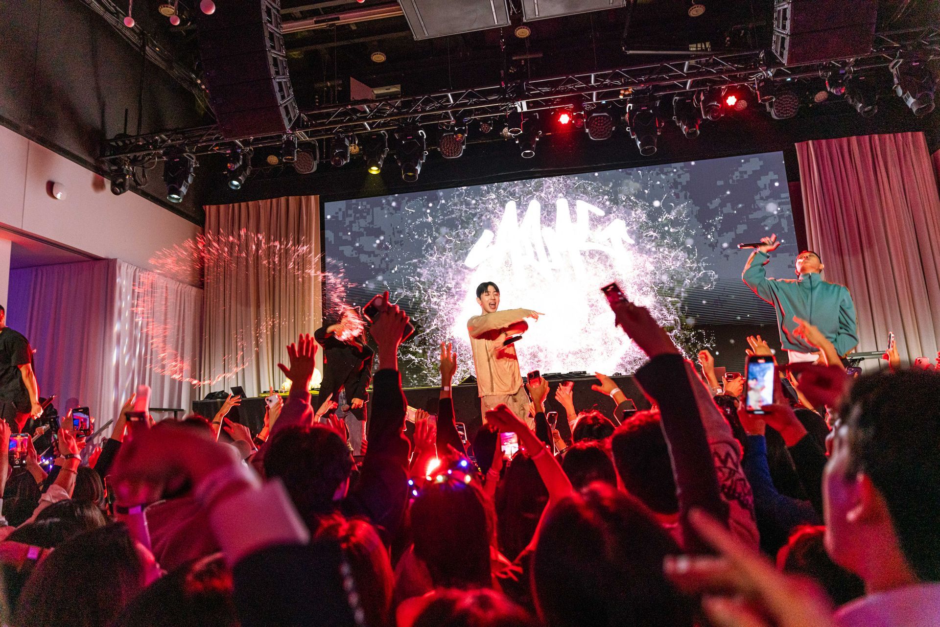 Concert crowd with performers on stage with a large screen displaying graphics. Red stage lights. People raise phones.