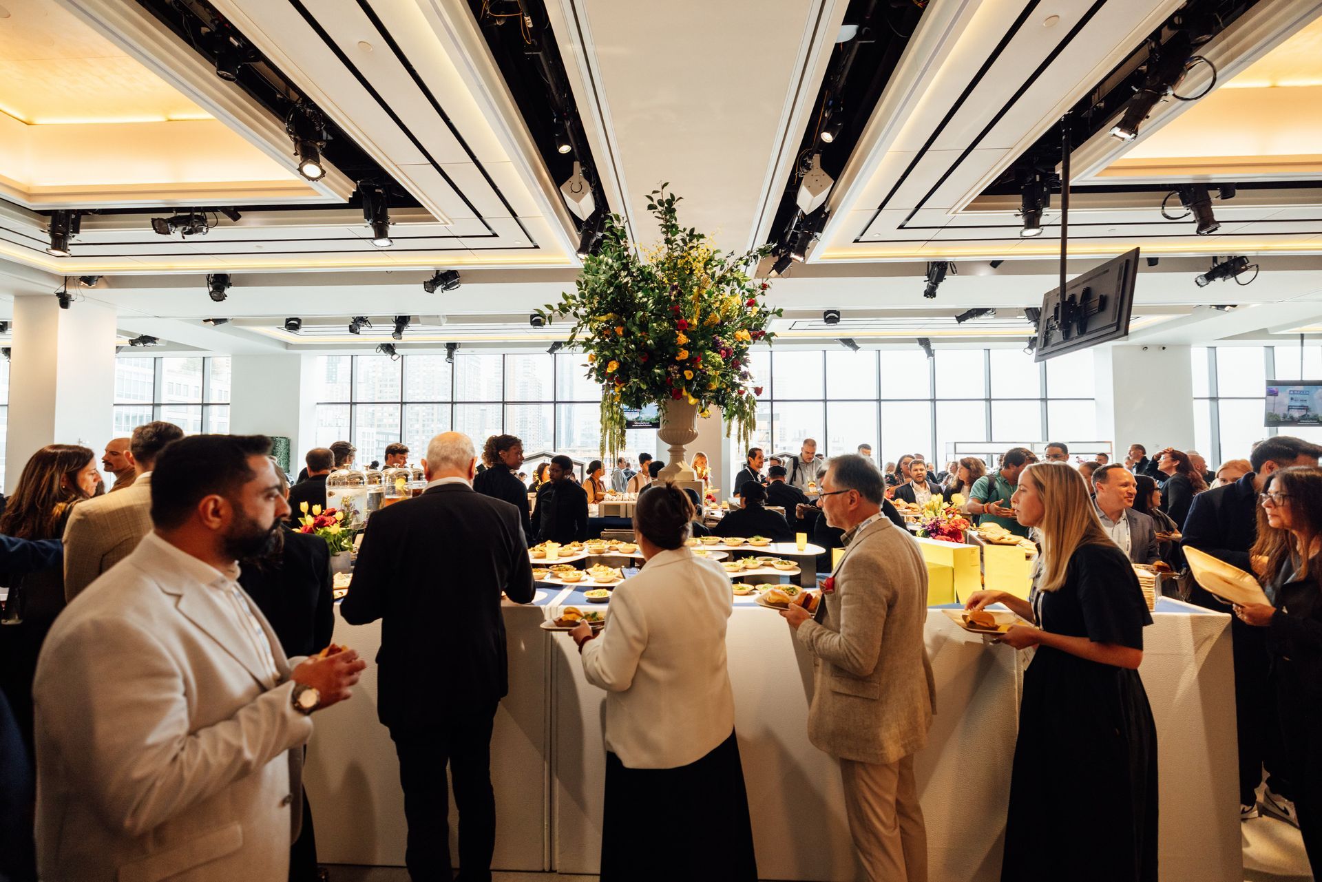 People at a reception, standing and mingling. Tables with food and a floral centerpiece. 