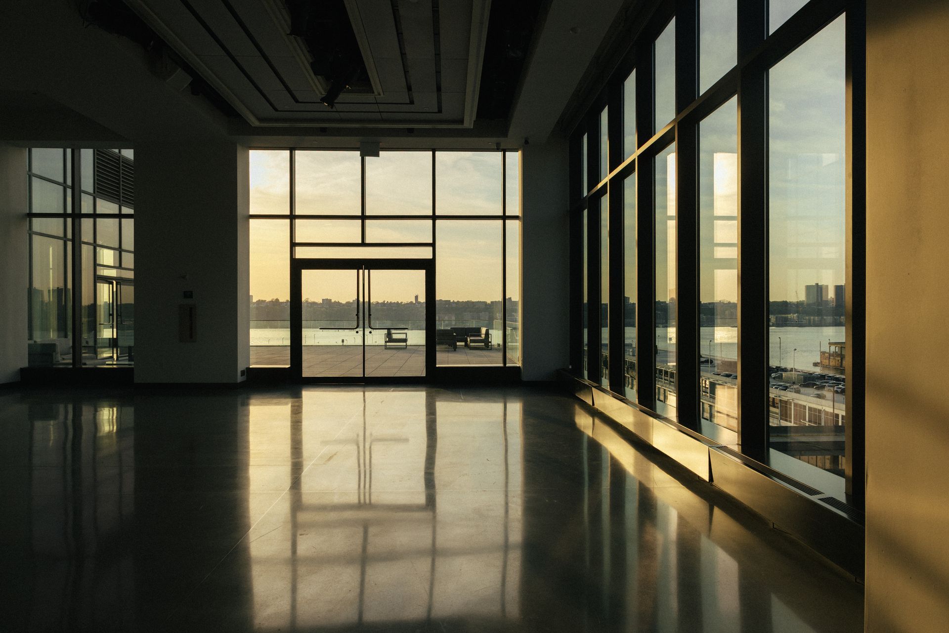 Empty modern interior with large windows, reflecting sunlight and overlooking a waterfront view.
