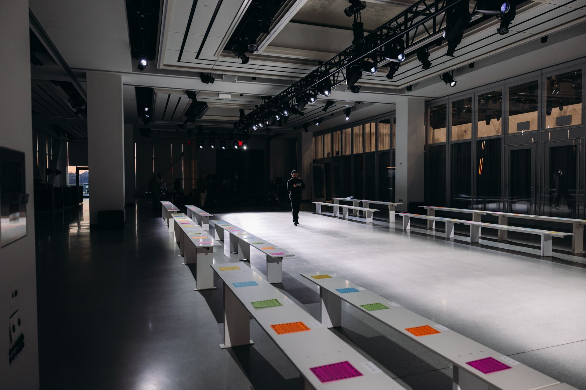 Empty fashion runway with rows of benches; a person stands on the runway under the lights.