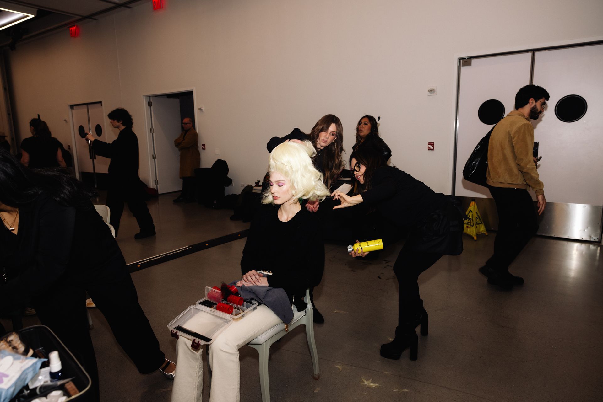 People in a bright room, one person has blonde hair being styled. Others are walking around, some with cameras.