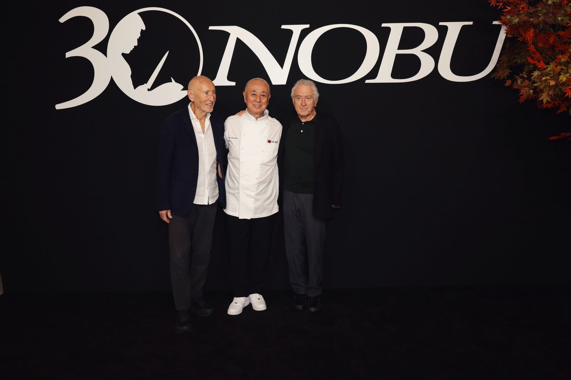 Three men posing in front of a “Nobu” logo. One in chef whites, the others in dark suits. Black backdrop.