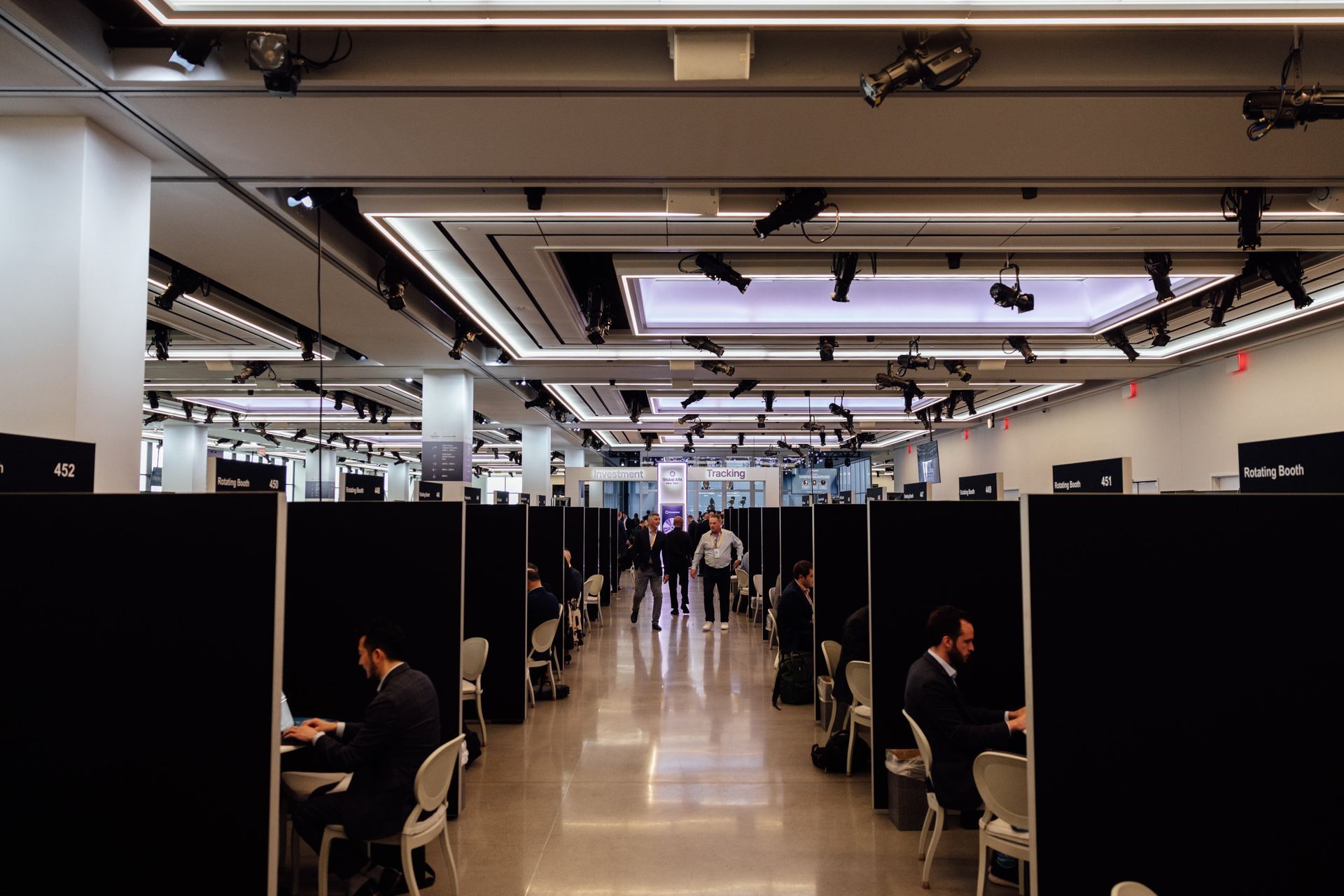 People working at computers in a long, well-lit room with individual black partitions.