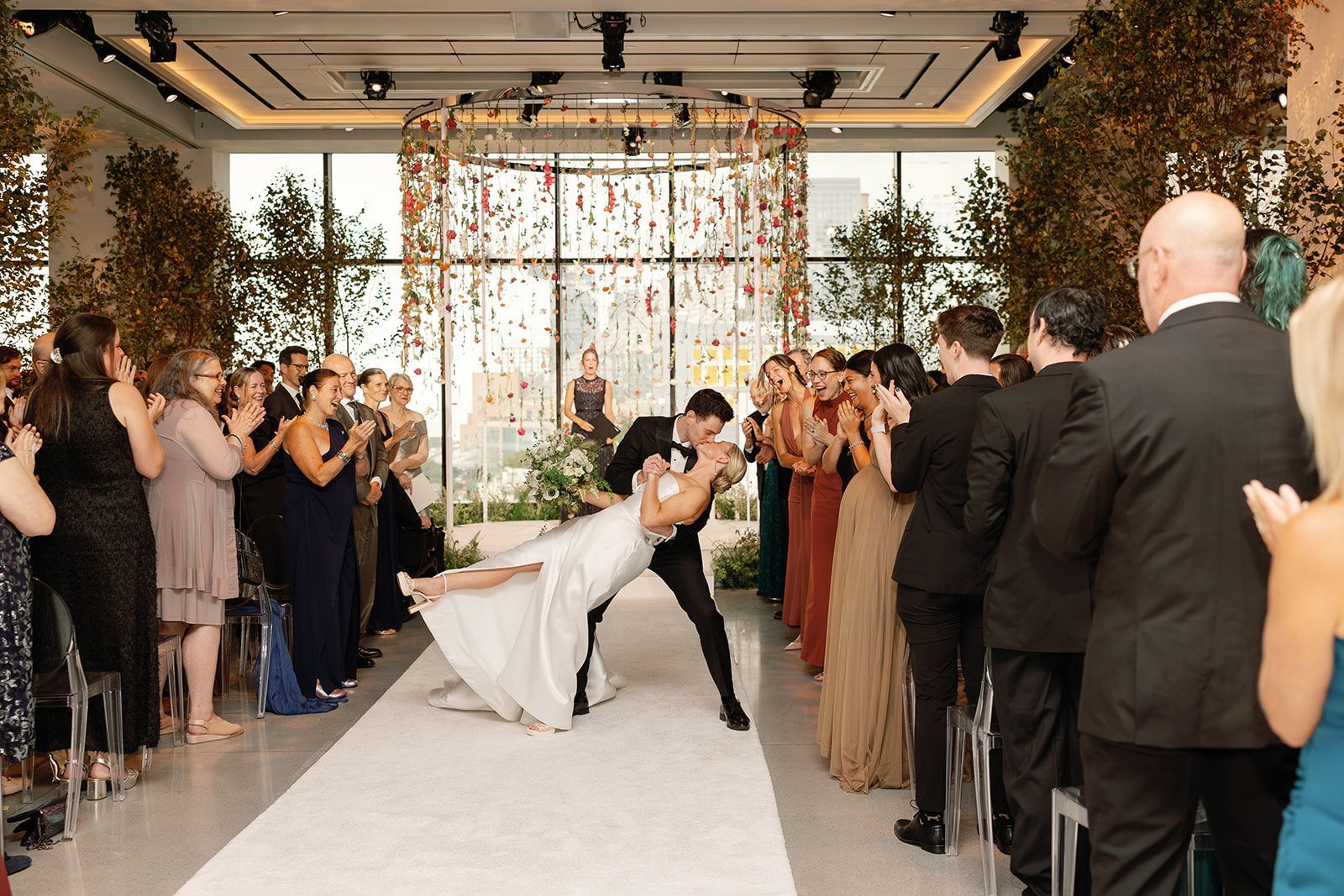 Bride and groom kiss at the end of a white aisle as guests applaud at a wedding reception