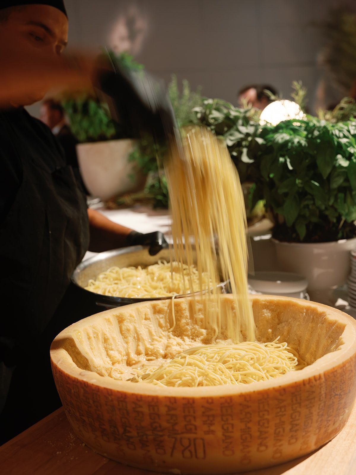 Person pouring noodles into a wooden bowl at a table with potted plants nearby