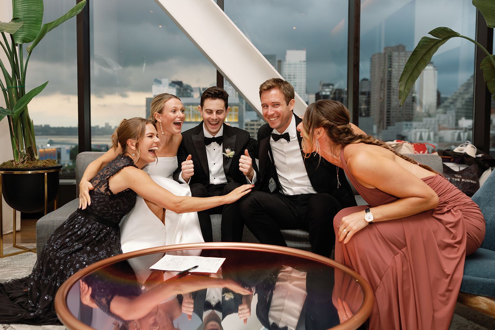 Five people in formalwear laughing and posing on a rooftop lounge with city views.