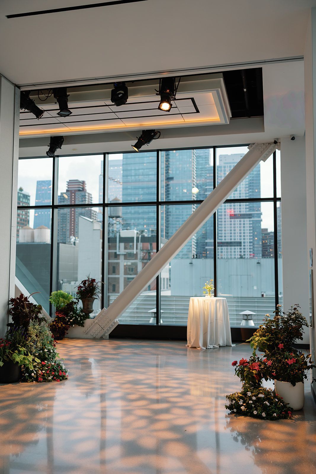 Empty event space with floor-to-ceiling windows, city view, spotlighted floor, and floral arrangements.