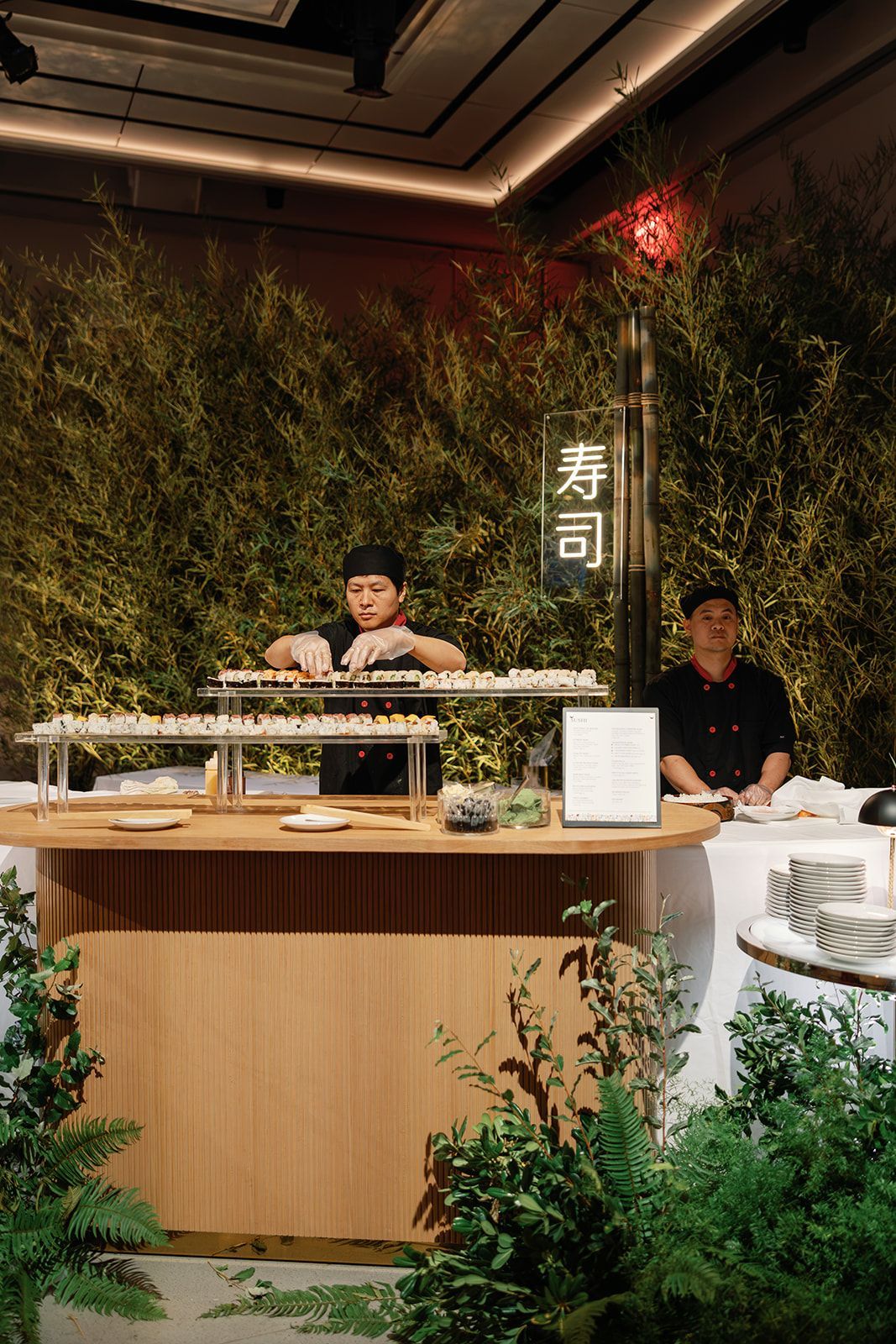 Sushi bar counter with two chefs behind a wooden station, framed by lush green bamboo decor.