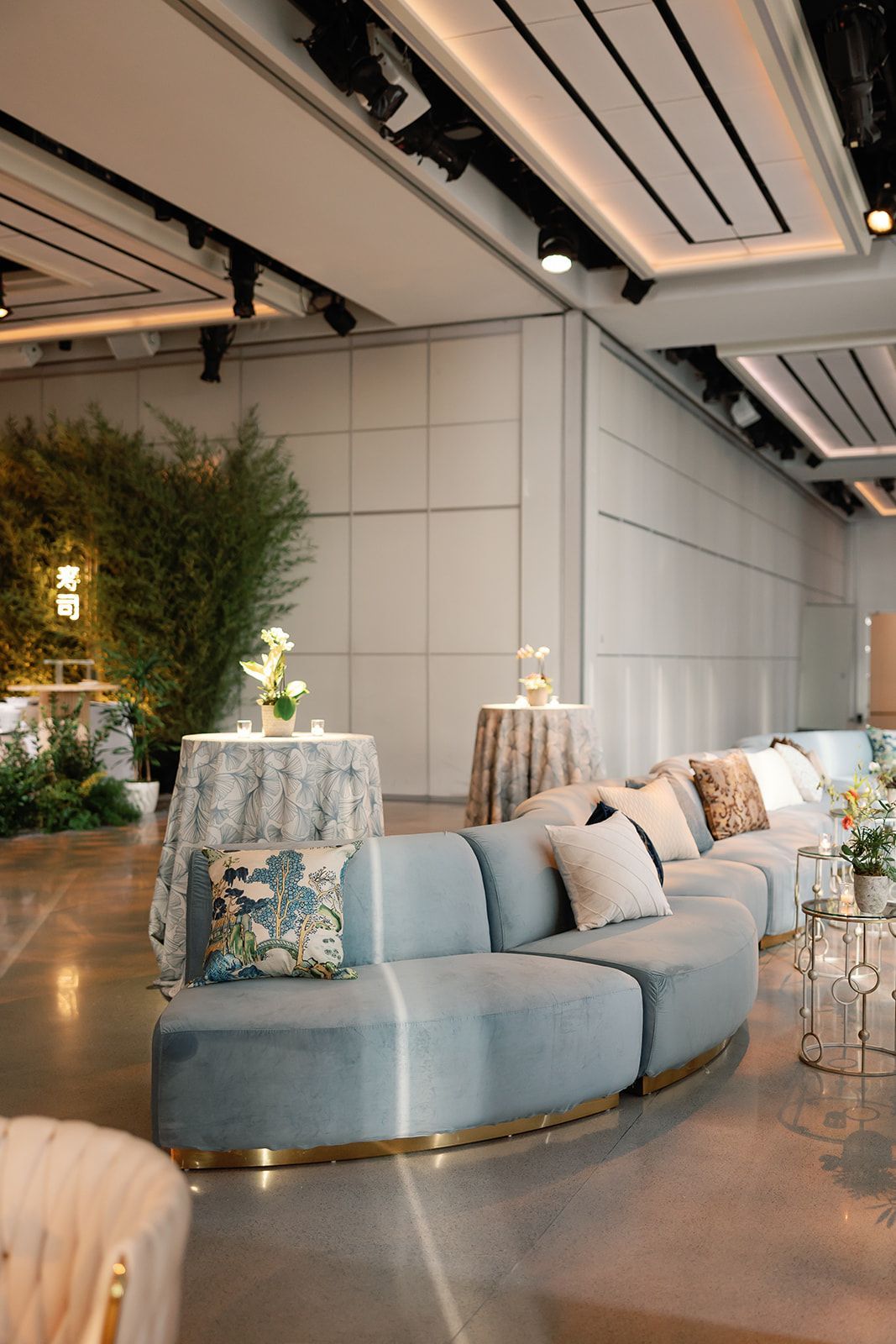 Modern lounge with blue curved sofa, floral pillows, tables, plants, and ceiling spotlights