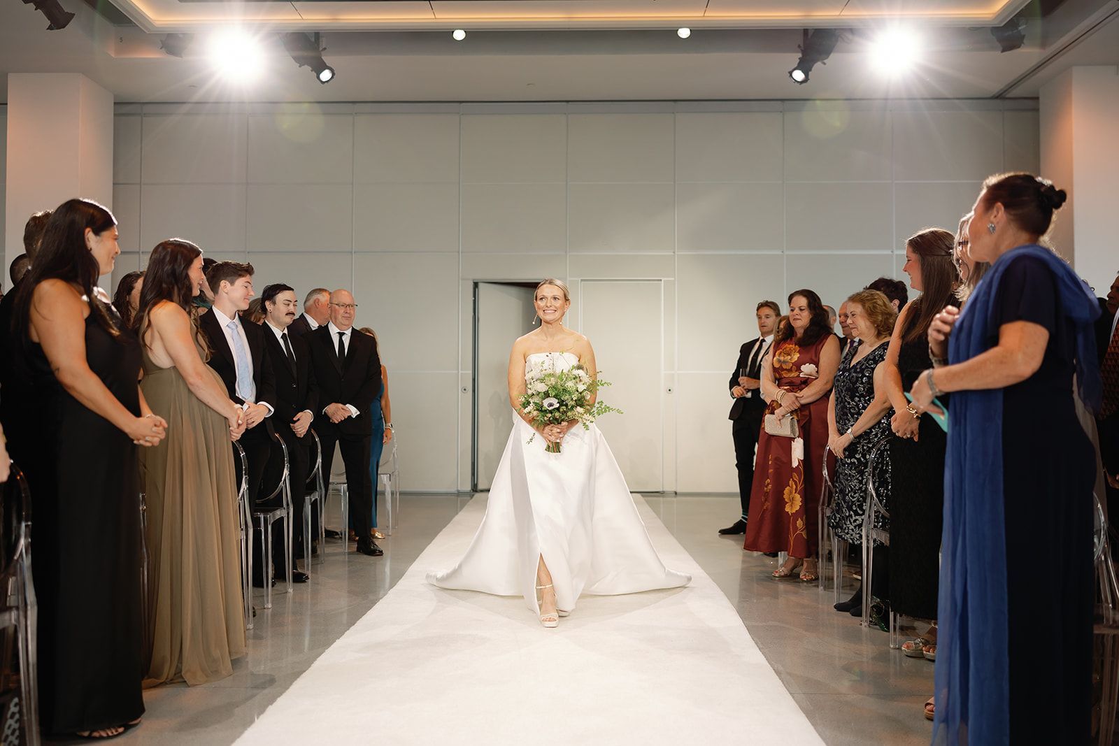 Bride walking down aisle between guests, holding bouquet in a bright indoor wedding venue